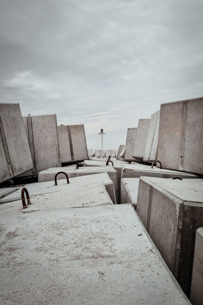 Concrete blocks on the shore guide the way to a distant lighthouse under a cloudy sky.